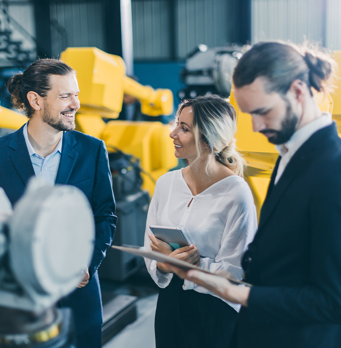 A customer and two sales reps, standing in front of yellow industrial machinery, with one person holding a tablet.