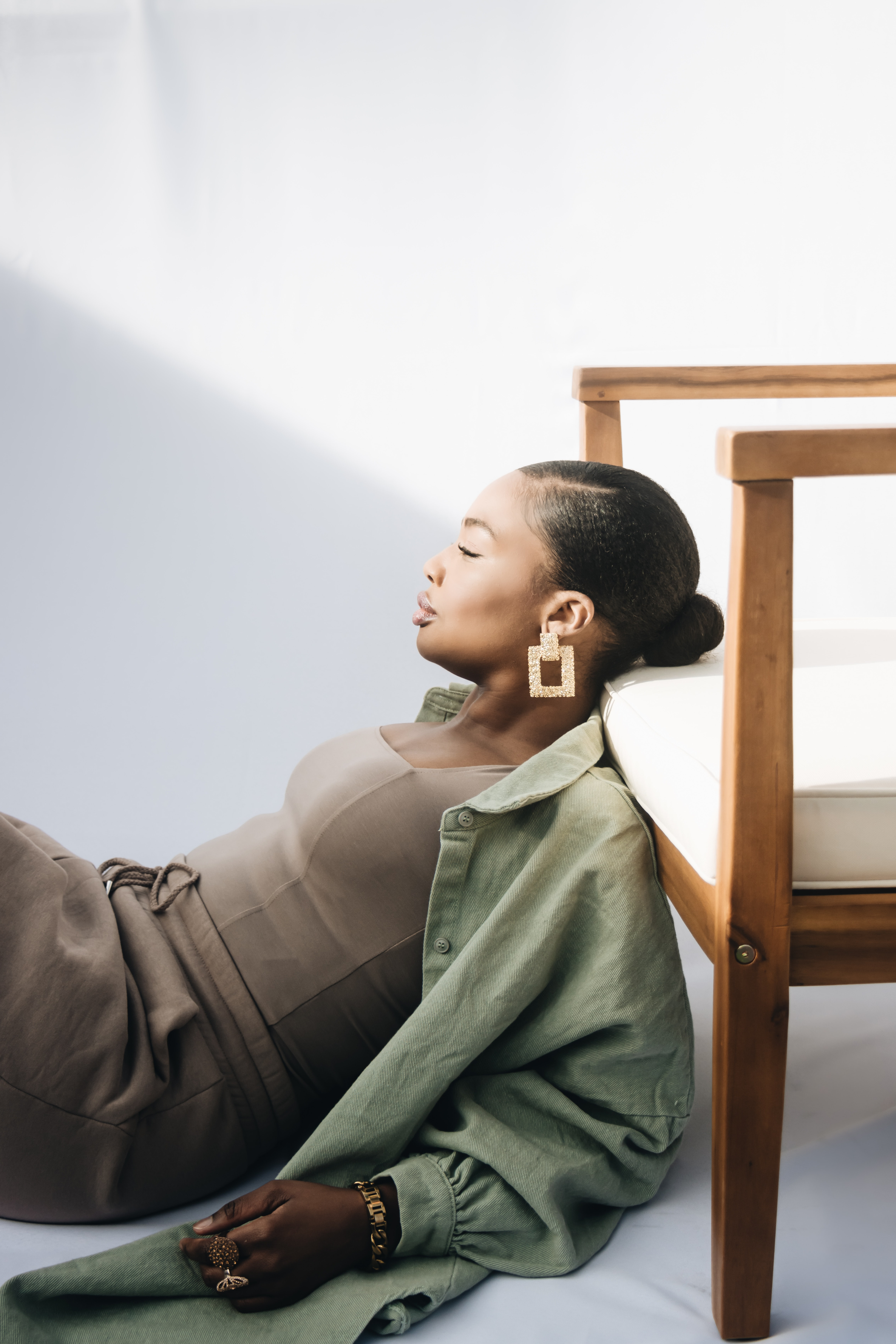 Fashionable woman with sleek hair, wearing large geometric earrings, leaning against a wooden chair in soft natural light.